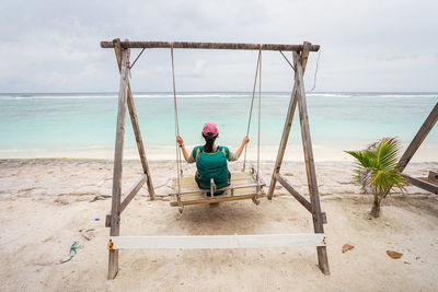 Back view of unrecognizable female traveler sitting on swings and enjoying fresh breeze while resting during summer holidays on sandy beach of hulumale island in maldives