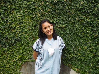 Portrait of smiling young woman standing against ivy wall