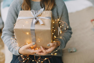 Midsection of young woman holding christmas present with illuminated string lights at home