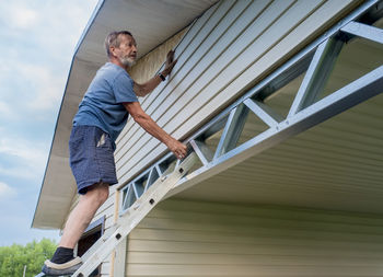 Full length of mature man standing outside house on ladder