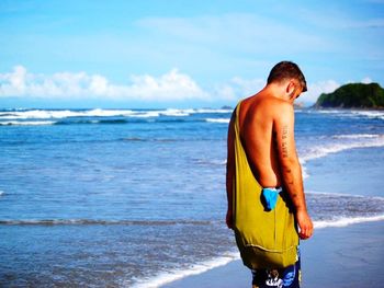 Shirtless man with shoulder bag walking on shore at beach