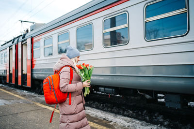 Girl on the platform of the station with a red backpack and a bouquet of tulip flowers