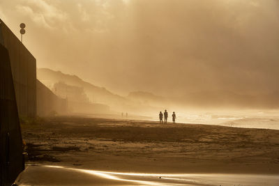 Silhouette people walking at beach against moody orange sky during sunrise