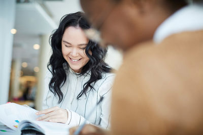 Smiling woman reading book while studying with friend at university cafeteria