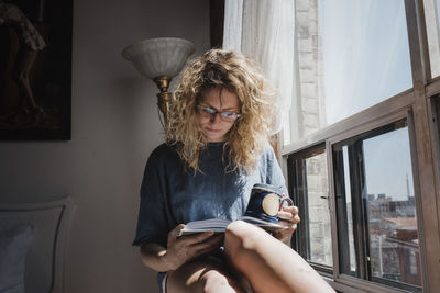 Woman holding cup while reading book on window sill at home