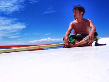 Full length of shirtless man sitting against blue sky