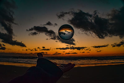 Person hand by sea against sky during sunset