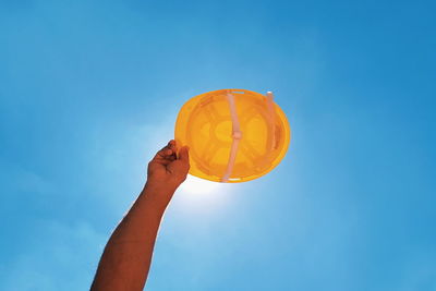 Low angle view of hand holding hardhat against clear blue sky