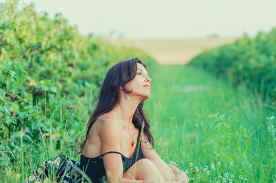 Beautiful woman with black hair sitting on grass near currant bushes. hand on knee, bare shoulder.