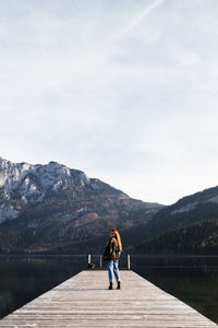 Back view of lonely female traveler standing on wooden pier near lake with snowy rocky mountains on shore in sunny autumn day