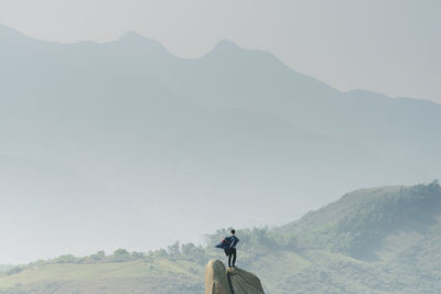 Rear view of man standing on rock against mountains in foggy weather