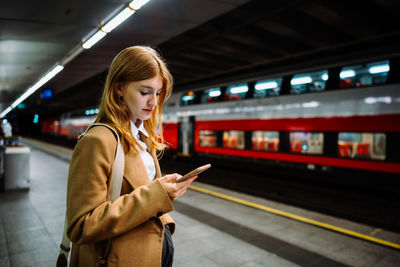 Young woman stands at a subway station and looks at her mobile phone