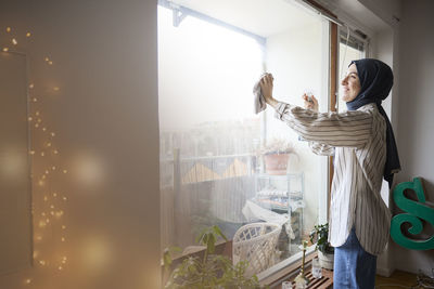 Happy woman in headscarf cleaning windows at home