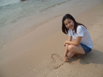Portrait of smiling woman making heart shape while crouching at beach
