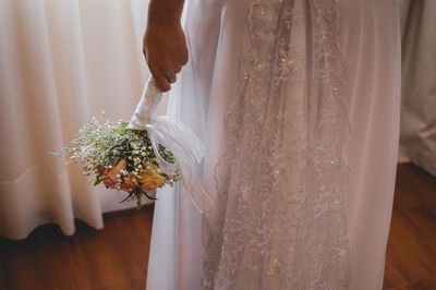 Midsection of bride holding flower bouquet while standing at home