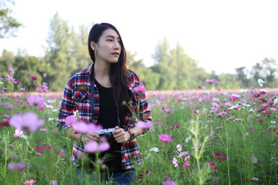 Thoughtful young woman looking away while holding camera amidst flowering plants