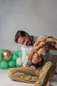 Father playing with small baby while holding legs and tickling in studio decorated with balloons for celebrating birthday together