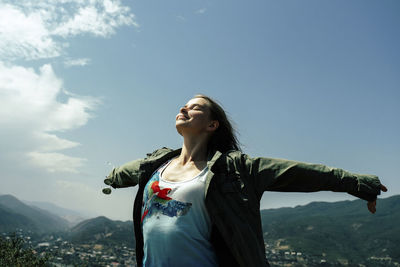 Happy woman with arms outstretched standing on mountain against blue sky during sunny day