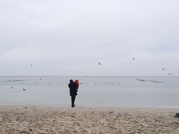 Rear view of mother carrying baby at beach against sky