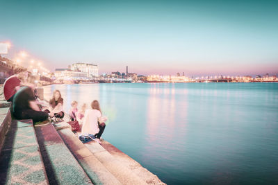 Friends sitting on steps by river in city against sky during sunset