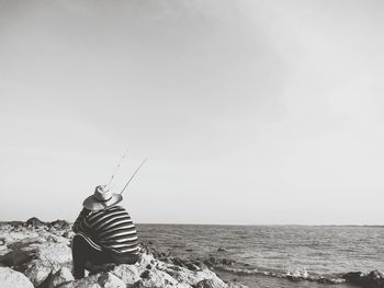 Rear view of man sitting on shore by sea against sky