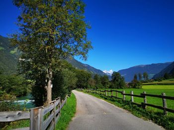 Empty road along plants and mountains against clear blue sky