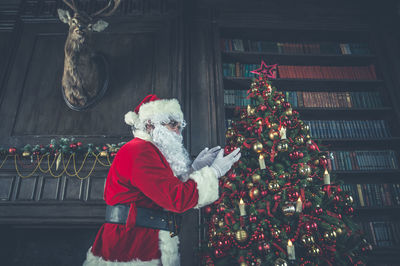 Side view of man wearing santa claus costume standing by christmas tree at home