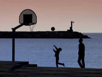 Silhouette men with ball on shore against sky during sunset