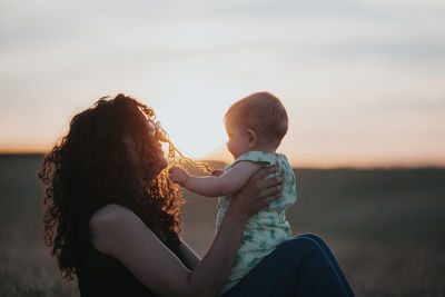 Side view of woman with baby sitting on field during sunset