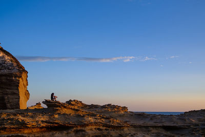 Woman sitting on rock formation against sky during sunset