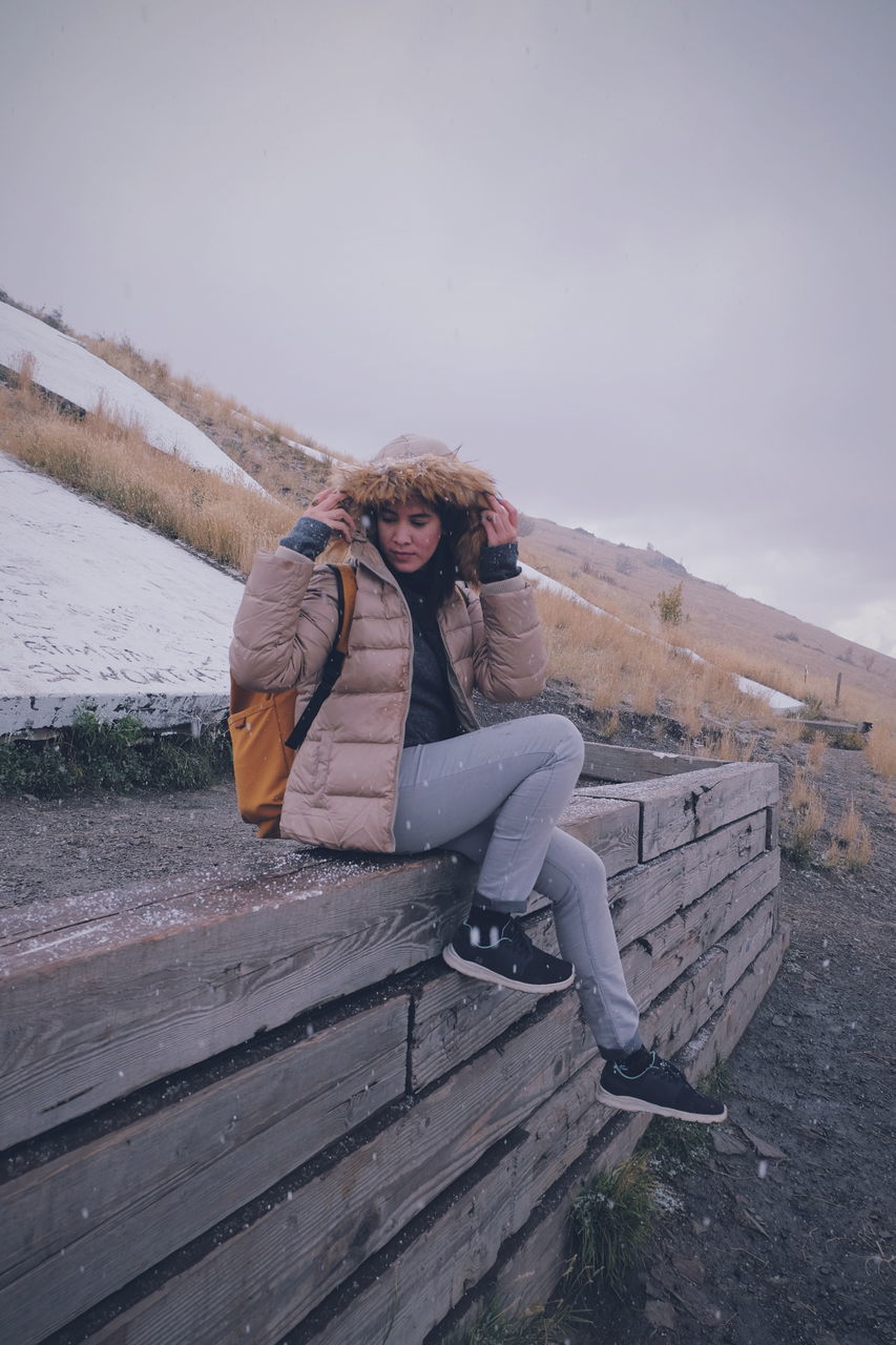 winter, one person, leisure activity, real people, lifestyles, full length, warm clothing, sky, cold temperature, young adult, clothing, front view, sitting, wood - material, bench, nature, looking at camera, young women, outdoors, scarf