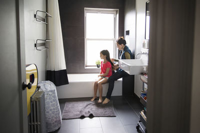 Mother tying hair of daughter while sitting in bathroom