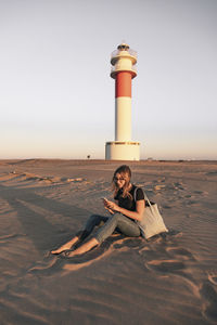 Woman using smart phone sitting on sand in front of lighthouse at beach