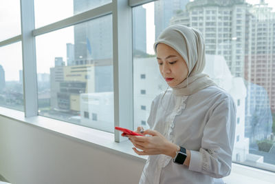 Young woman looking through mobile phone while standing on window