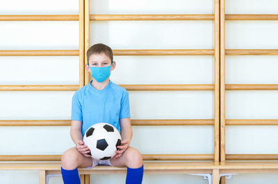 Portrait of boy wearing mask with soccer ball sitting on bench