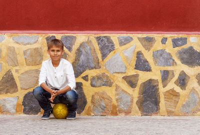 Portrait of smiling boy sitting on ball by wall