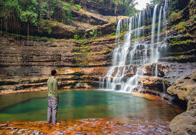 Young man standing near waterfall flowing streams falling from mountain at morning 