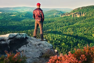 Tall man in black with red baseball cap on sharp cliff and watch to valley. colorful summer morning