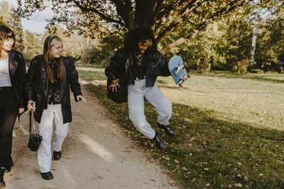 Cheerful woman jumping with skateboard while female friends walking by in park