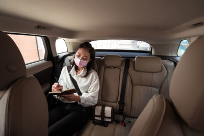 Female in formal wear in protective mask writing in notepad while riding on passenger backseat in comfortable cab