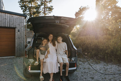 Portrait of smiling woman sitting with sons in electric car trunk on driveway