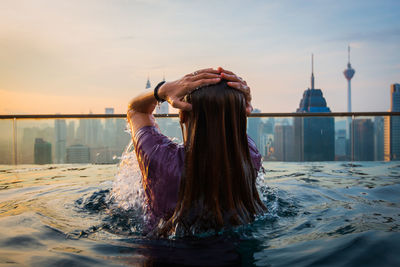 Rear view of woman swimming in infinity pool against city