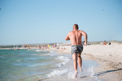 Full length of shirtless man running on beach against clear sky