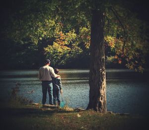 Rear view of father and son standing by tree at lakeshore