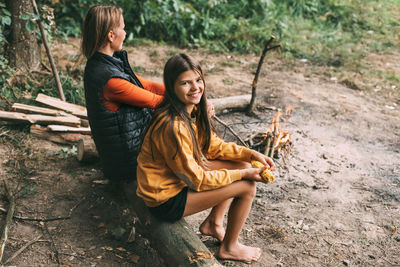 A mother with her teenage daughter is sitting on a tree near a campfire in the forest 