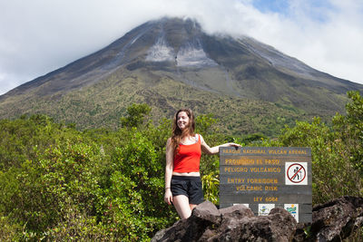 Smiling young woman standing by sign against volcano