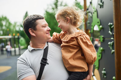 Smiling father embracing cute daughter on playground in summer while looking at each other