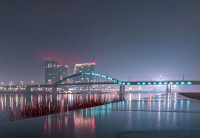 Illuminated bridge over river by buildings against sky at night