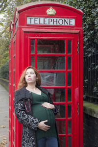 Thoughtful pregnant woman leaning on telephone booth at sidewalk
