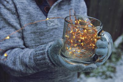 Midsection of person holding illuminated string lights in glass mug during winter